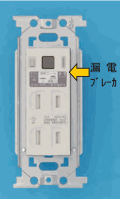 取付枠に付いた白い縦長の器具で、上部にボタンと表示があり、黄色の矢印と 漏電 ブレーカ の字が添えてある。下部に縦長の刃受2つの組が上下に2組ある