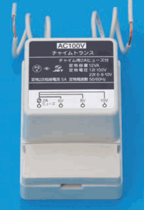 灰色の箱形の器具で、上から白い線が出ている。正面に AC100V、チャイムトランス、チャイム用2Aヒューズ付、定格容量 12VA、定格電圧 1次 100V 2次 6-8-10V、定格2次短絡電流 5A、定格周波数 50/60Hz の字とPSE・JETのマーク、下部に 2A ヒューズ・6V・8V・10V の字のある端子がある