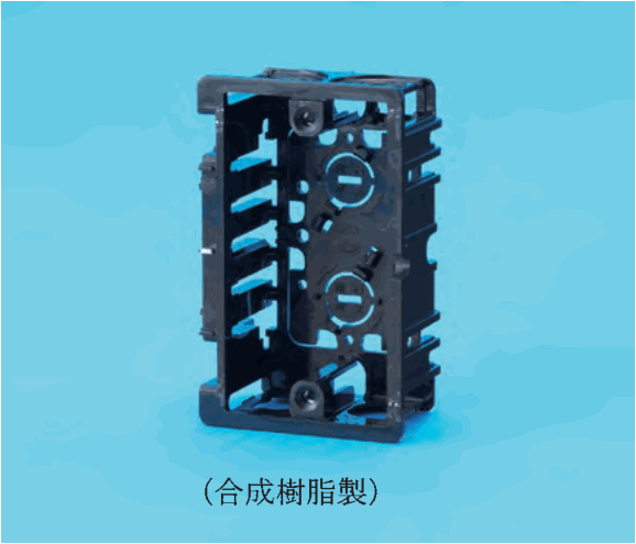 水色の背景に置かれた黒色の樹脂製の縦長の箱形の材料。前面が開いていて,上下に取付け用のねじ穴があり,奥の面に中央に横長の溝のある丸い部分が上下に2つある。側面には多数の細長いすき間がある。下に (合成樹脂製) の字がある