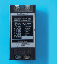 黒い箱形の器具で,上下に2つずつ端子ねじがあり,上に 100V,下に 24V の字。正面の表示に 小型リモコントランス(F型)(分電盤用)品番 WR2301,PSEとJETの表示,一次電圧 100V AC,周波数 50/60Hz,二次電圧 24V AC,二次電流 1.5A,定格容量 36VA,ヒューズ容量 2A,分電盤用,Made in Japan,ヒューズ容量2A の字がある