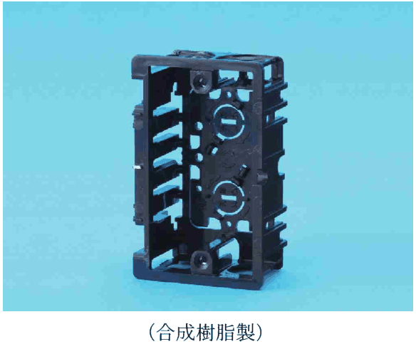 青い背景に置かれた黒色の長方形の枠状の材料。側面に多数の切り込みのある板が並び,奥の面に上下2つの丸い部分があり,上下の縁にねじ穴がある。写真の下に(合成樹脂製)の字