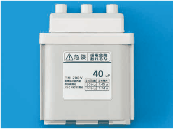 青い背景に置かれた灰色の箱形の機器。上部に3つの円筒形の突起がある。正面のラベルに 危険 感電危険 触れるな,40 μF,三相 200 V,50 Hz 1.45 A,60 Hz 1.74 A,JIS C 4901に適合 などの字がある